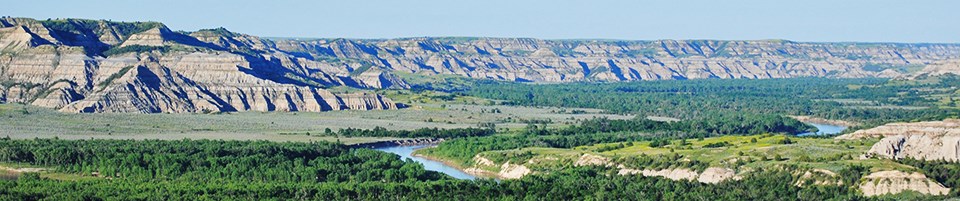 Tan and grey stripped buttes rise up from a wide green valley with a blue, winding river.