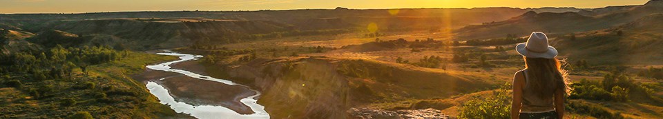A woman in a western style hat looks out across the Little Missouri River and badlands bathed in the sunset's golden light.