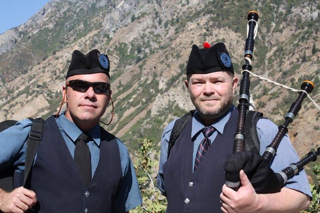 Two bagpipers stand in front of the mountain wearing kilts.