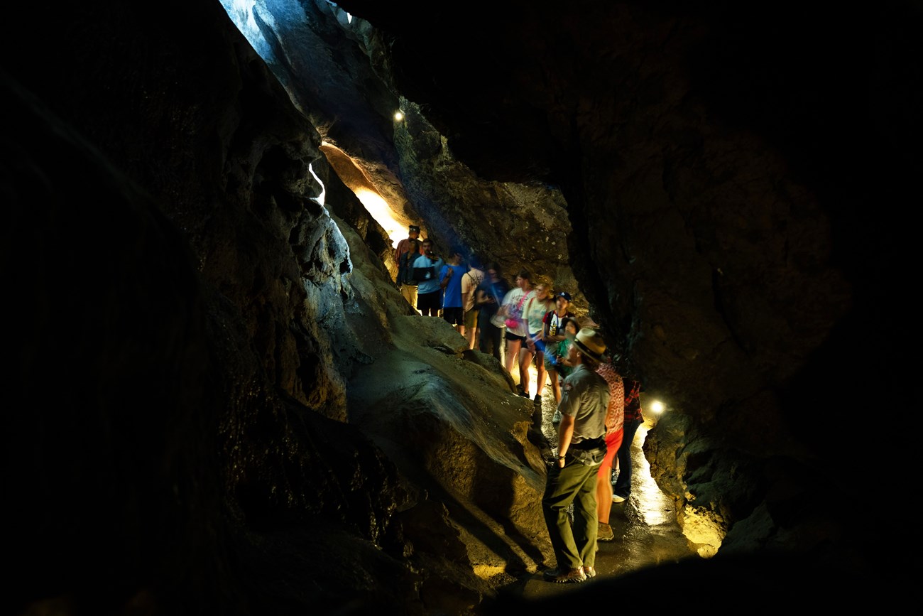 A ranger leads a group of 16 youth on a cave tour in a tight, dark passage. He shines a flashlight towards the ceiling as the group looks up.