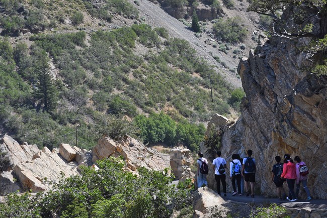 A group of 7 middle schoolers hike down a paved trail surrounded by rocky cliffs.