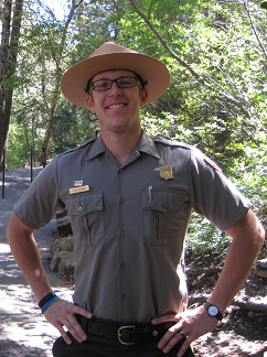 Ranger stands on paved hiking trail facing the camera with hands on hips. Green trees in background.