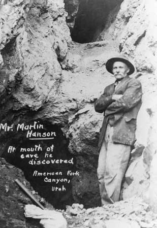 Martin Hansen stands at the mouth of the cave he discovered, Hansen Cave. The photo is in black and white, and Hansen is standing on a pile of rocks, just in front of the natural entrance.