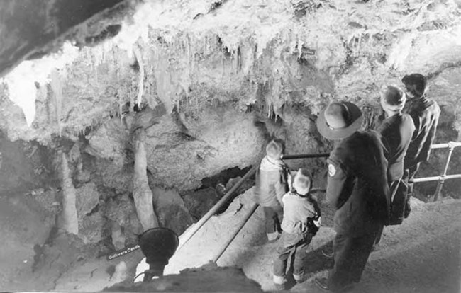 Two adults a two children looking at cave formations with a ranger standing a level above them. Image is black and white.
