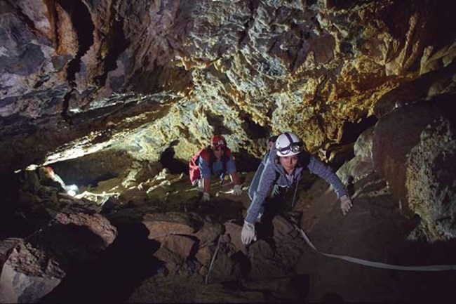 Two people wearing helmets and gloves climb up a steep slope in the cave.