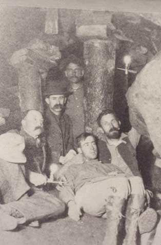 George Tyng with his hat on his knee and his two sons sitting beside each other.  CH Joy the photographer is sitting in the middle behind George.  One of George's miners is visible in the background by the candle reflection light. All the men are surrounded by what appear to be mining timbers inside of a rock tunnel.