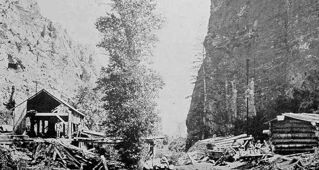 Black and white photo of an early sawmill in American Fork Canyon showing outbuildings and piles of logs.