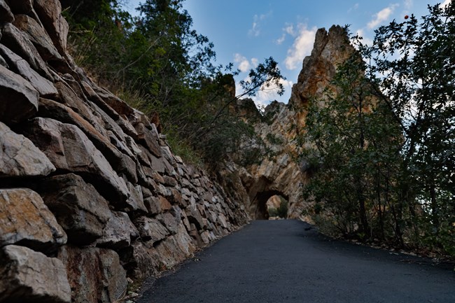 Rock wall on the left hand side of a paved path leading to a rock archway.