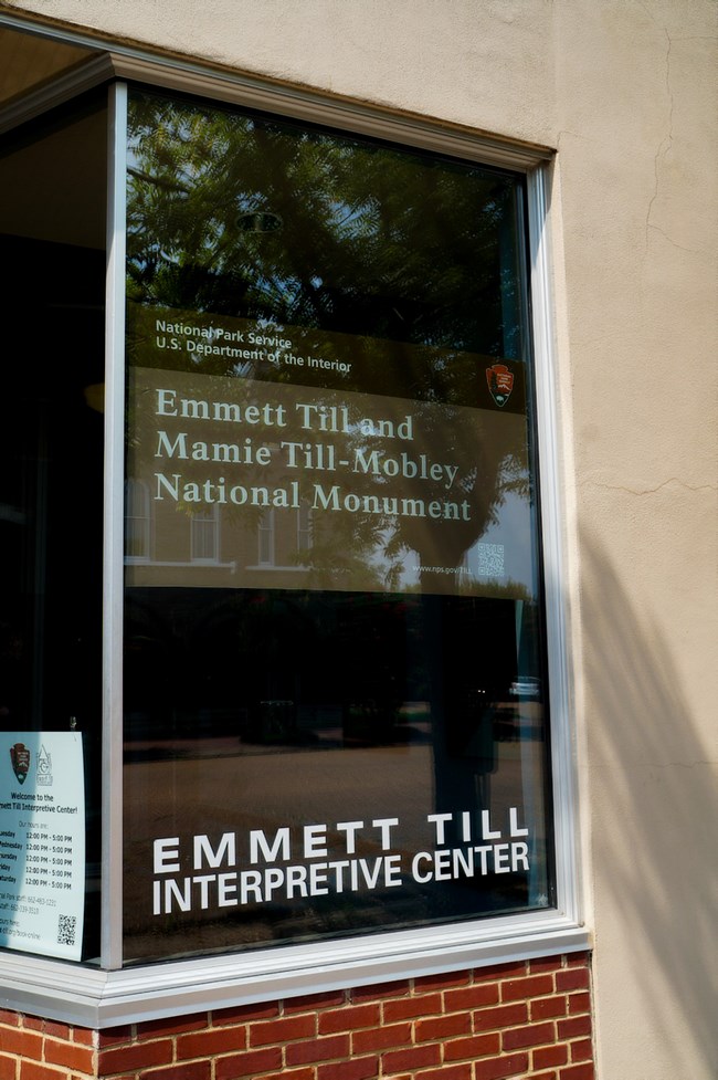 Building exterior focused on a window, where signs read "Emmett Till and Mamie Mobley Till National Monument" and "Emmett Till Interpretive Center"