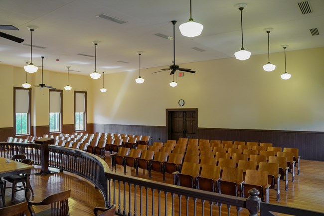Rows of stadium-style seats face the front of the room, where to wooden table sit, separated from the audience by a wooden banner