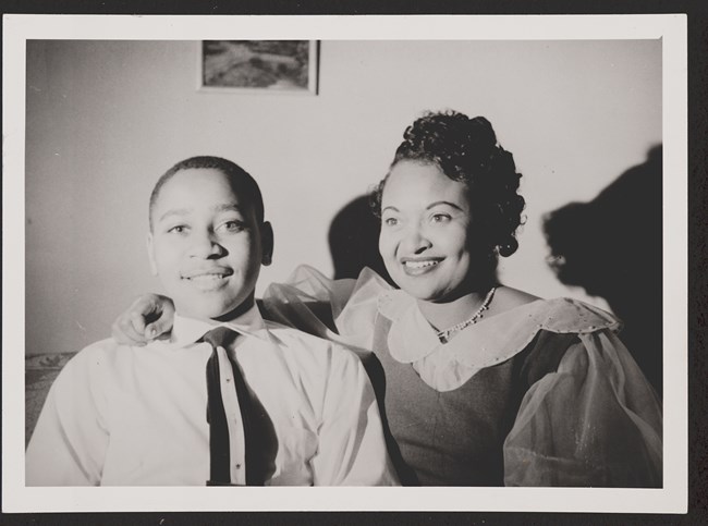 Two African-Americans sitting in a room and smiling toward the camera. A woman, on right, has her arm around a boy on the left.