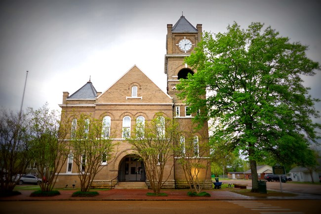 A two-story brick courthouse with a a triangular roof and clock tower on the right side.