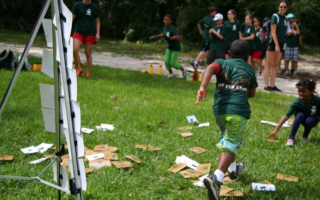 Image of students running back and forth on grass to construct a Timucuan Hut using a trifold display and cardboard. A group of students and teachers stands in back.