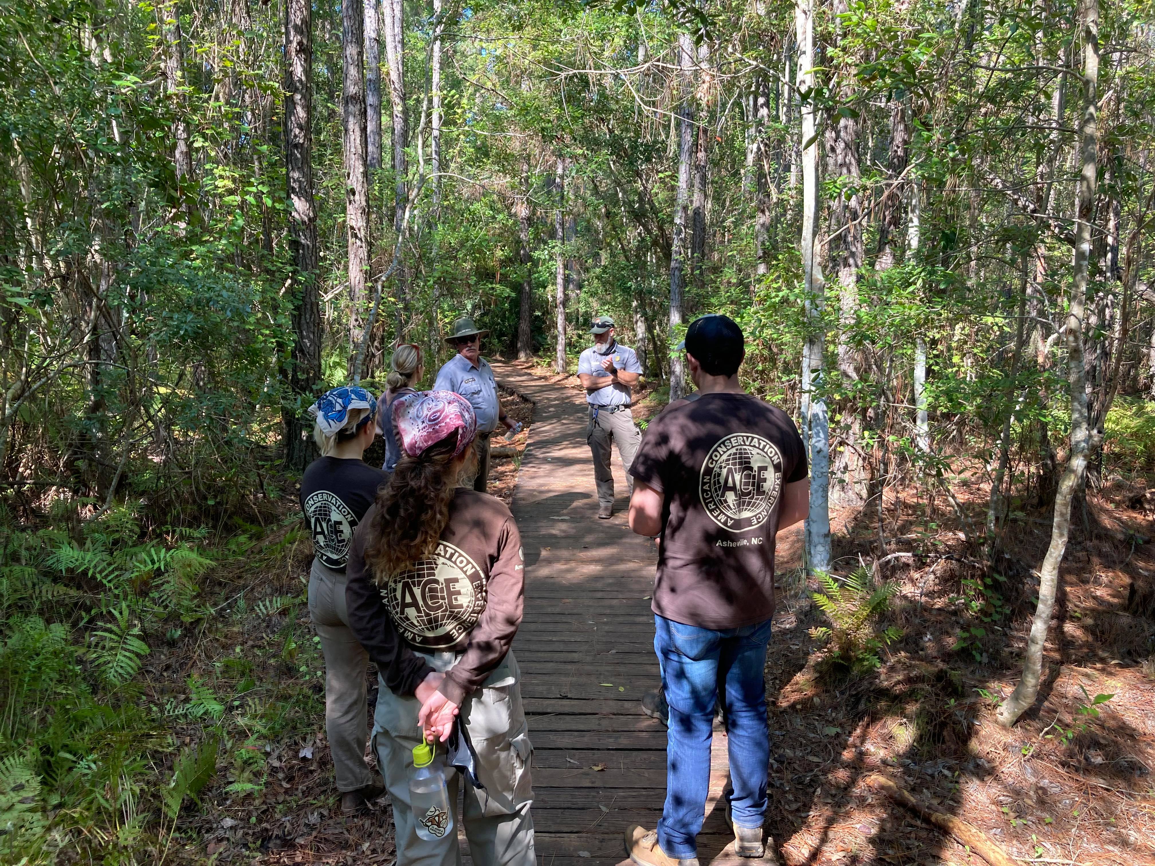 Rangers and ACE Meet on trail youth in brown ACE shirts talk to rangers on boardwalk in the woods