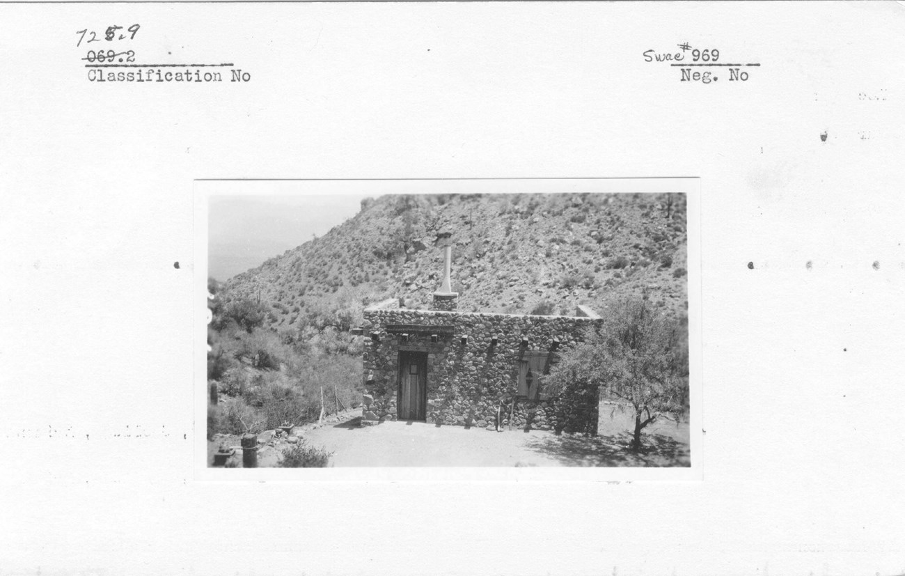 Black and white photo of a small stone structure with desert landscape around.