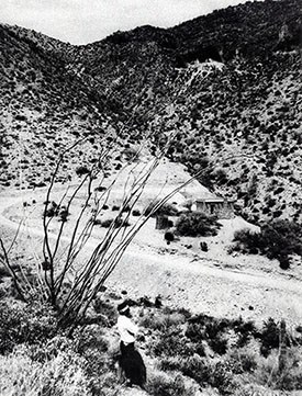 Black and white image of road leading in to Tonto National Monument with stone caretaker's house in distance.