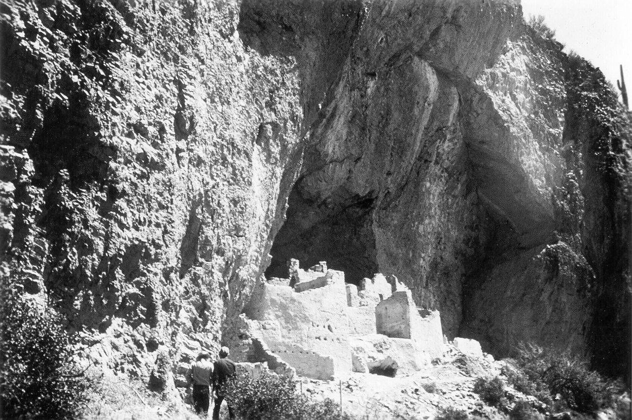 A black and white photo of two members of the CCC in front of the Upper Cliff Dwelling.