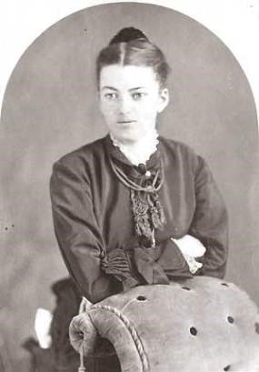 Black and white image of Angeline Mitchell standing while leaning on a chair in front of her.