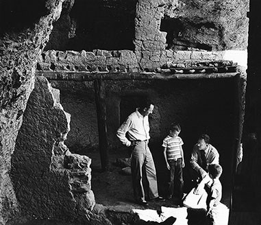 Charlie Steen kneels next to two children and one adult visitor in the Lower Cliff Dwelling. Partial ceiling and walls surround them.