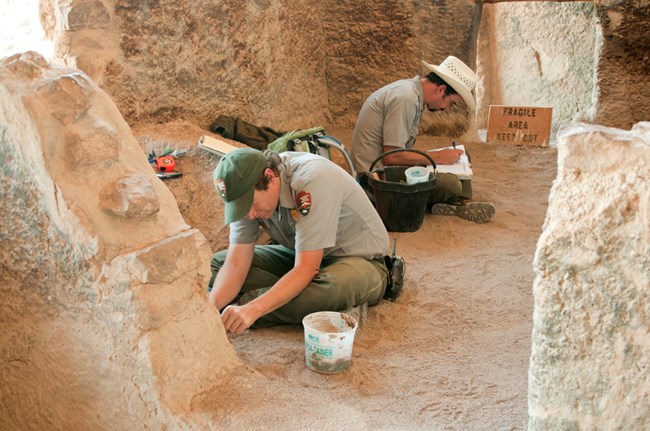 Two men in NPS uniform sit in a cliff dwelling. One works to stabilize a wall. The other writes on a clipboard.