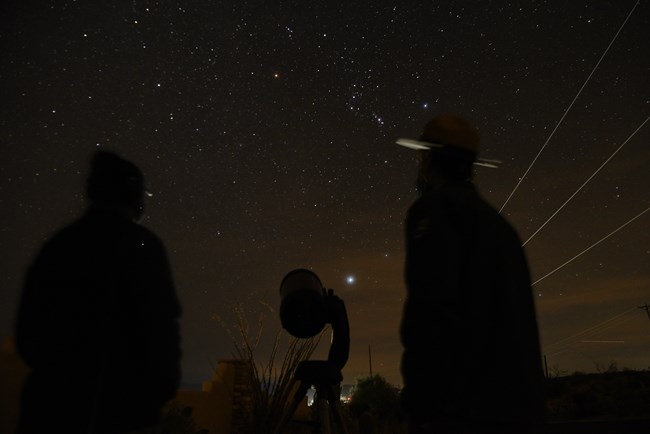 Two people stand with a telescope looking up at a star filled sky.