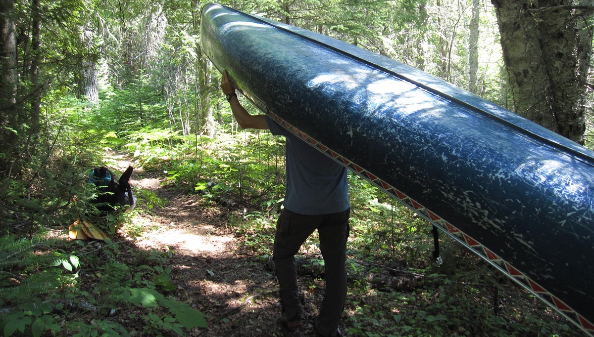 A person portages a canoe down a trail surrounded by forest towards a backpack and paddles on the ground.