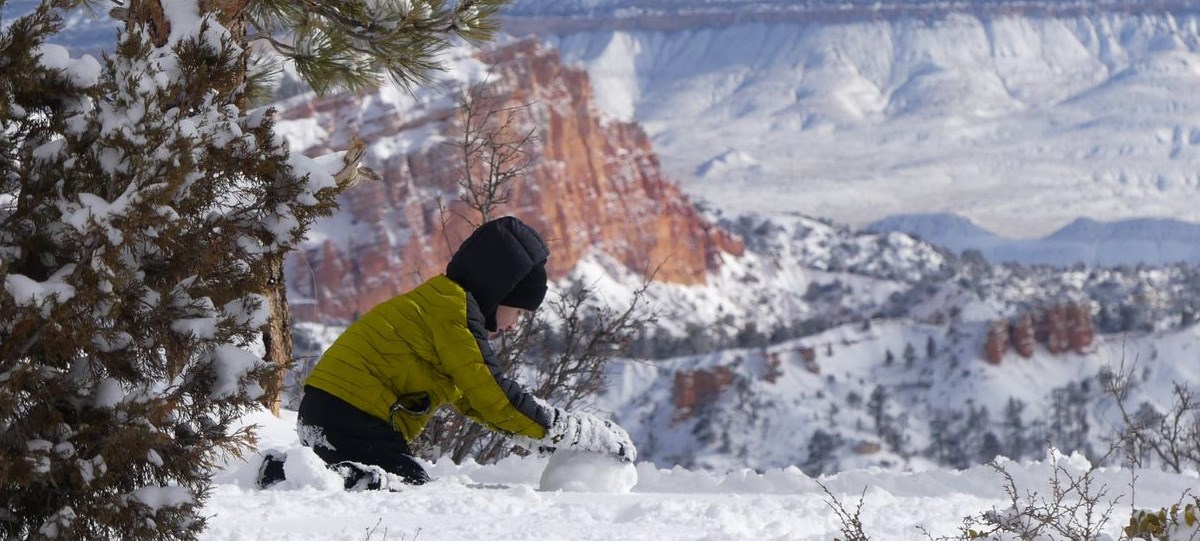 A young boy in a blue coat plays in the snow with snow covered rock formations in the background