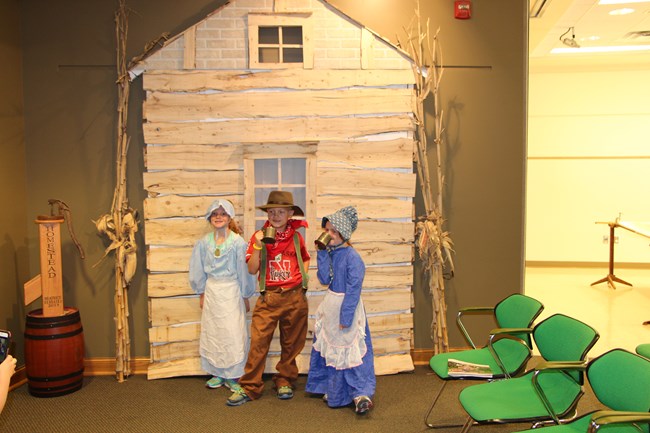 Three children wearing period piece costumes stand in front of a log cabin facade.