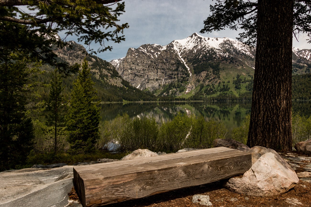 A wooden bench sits beside a lake surrounded by mountains.