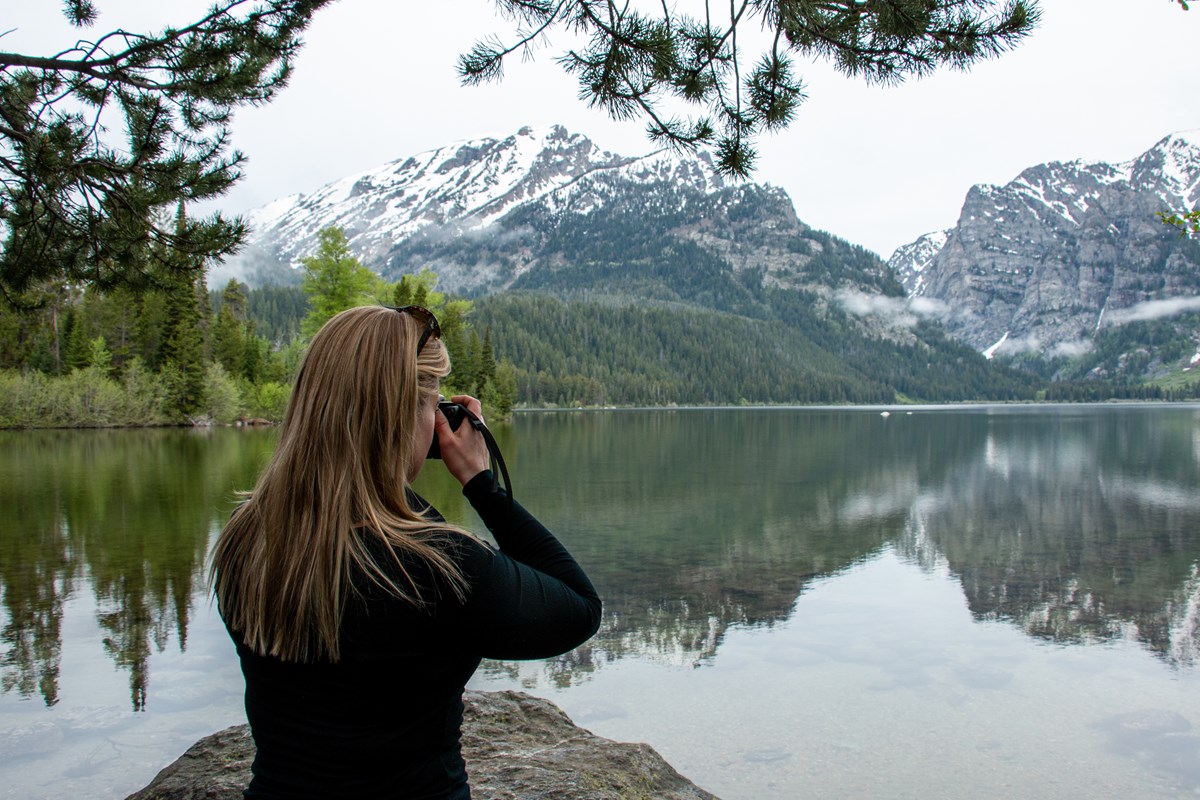 A woman takes a photo at a lakeshore.