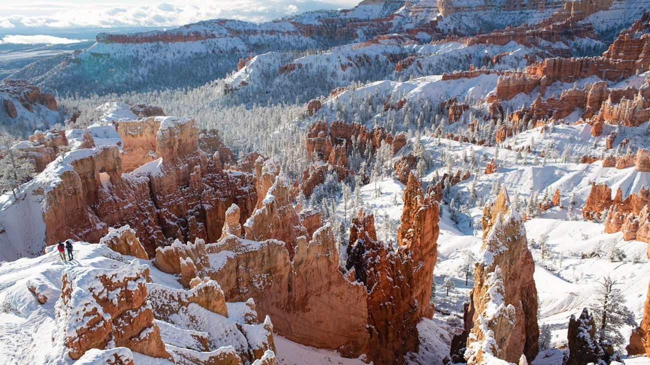 Two hikers walk amongst snow covered red rock formations in an amphitheater