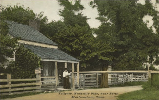 A historic image of a person standing in front of a small house next to a dirt road.