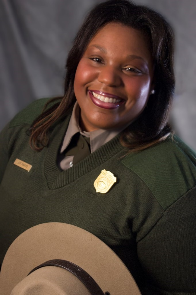 Dr. Joy G. Kinard, African American woman smiling in green National Park Service sweater holding a beige National Park Service hat