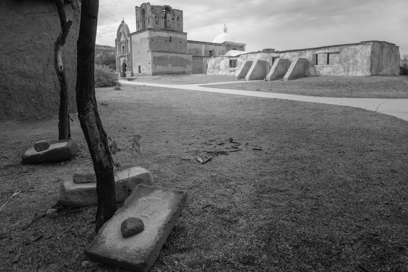 Black and white photograph of grinding stones on the ground with adobe church in background.
