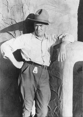 historic photo of man in jaunty ranger flat hat, leaning against earthen building