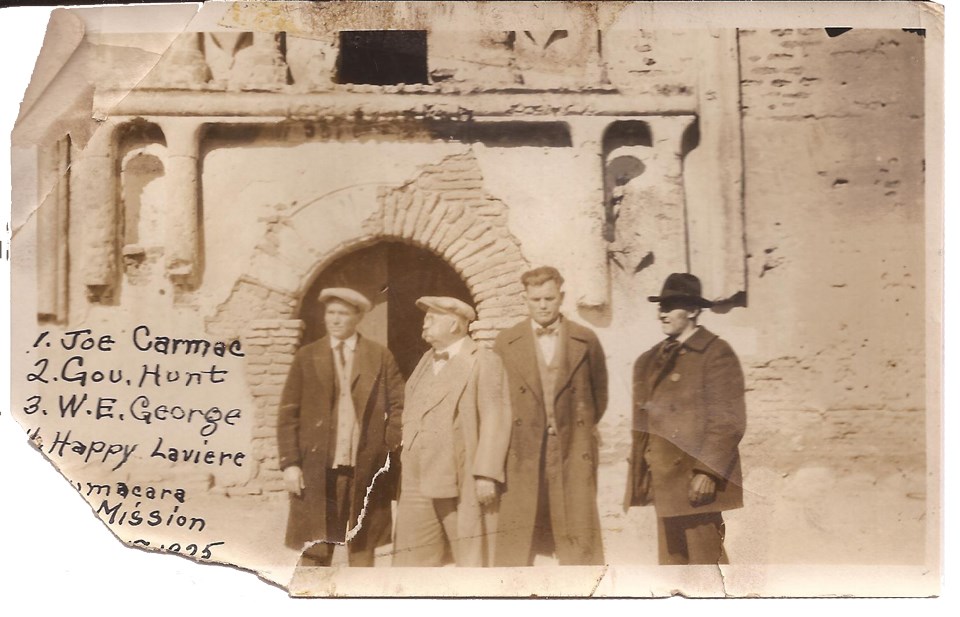 sepia-toned photograph with torn corner featuring four men in coats in front of church