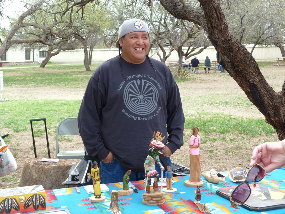Demonstrator stands at a table with finished carved and painted kachina figures.