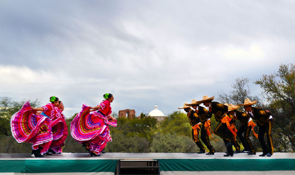 folklorico dancers on stage, men on right, women on left, church dome and trees in the middle, gray sky above