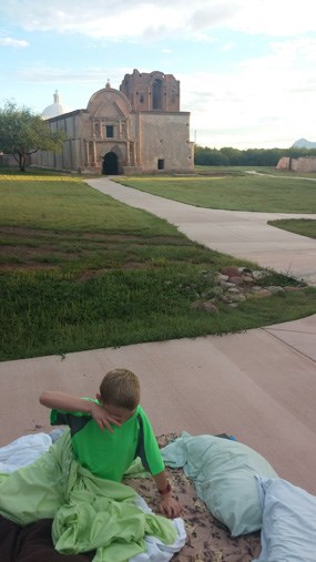 child rubbing eyes on bedroll in front of church at daylight