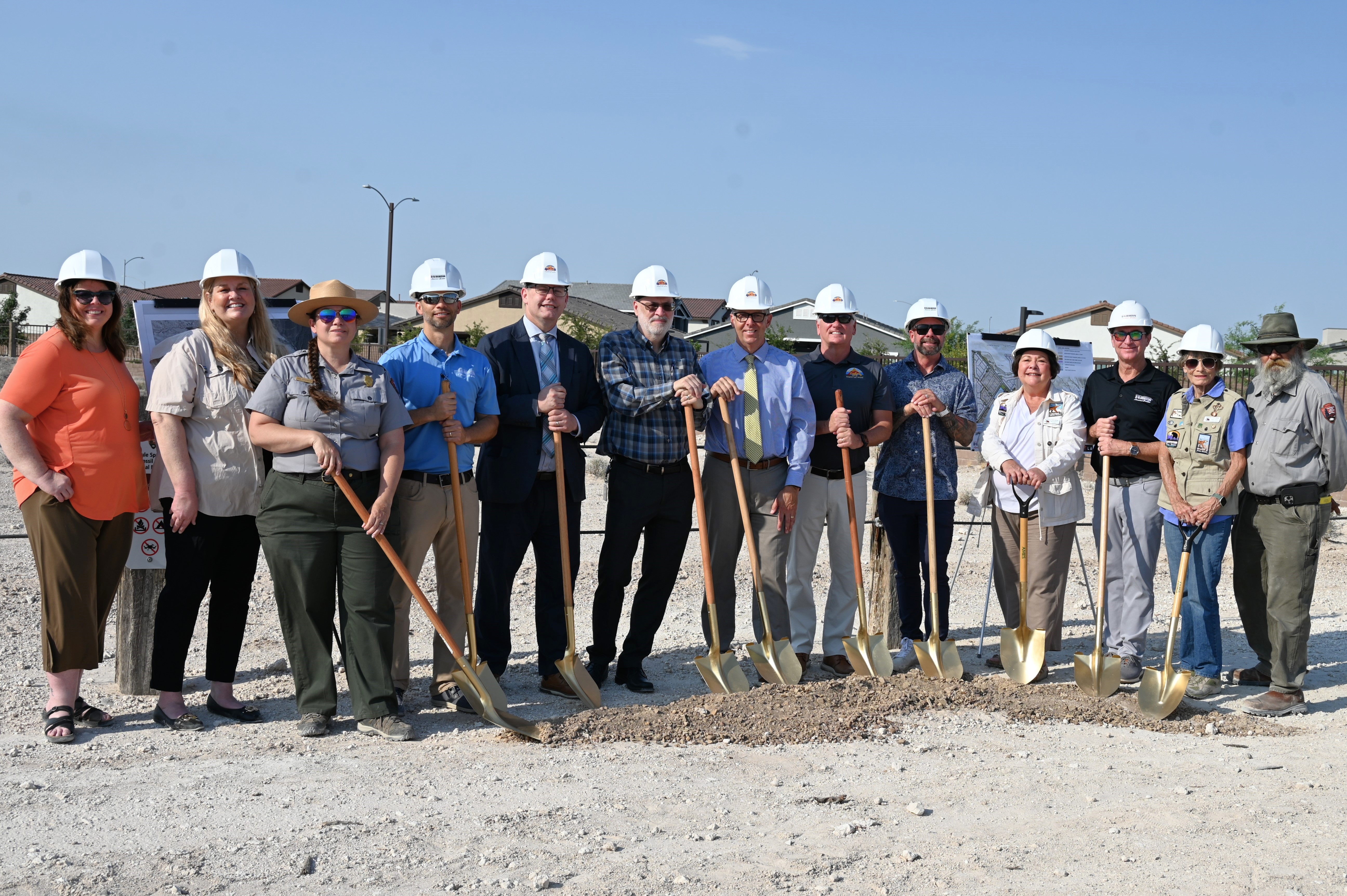 10 people wearing hardhats and 2 park rangers pose with shovels at a trailhead.