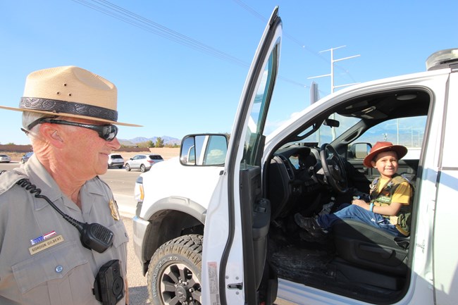 A park ranger standing next to a truck with a Junior Ranger sitting inside.