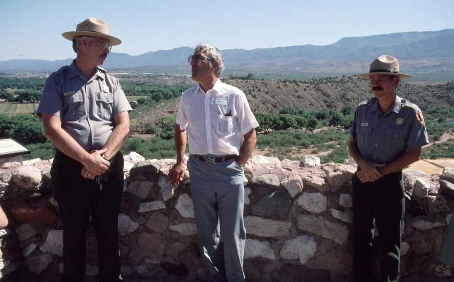 Two men in NPS uniform and one in a shirt and slacks stand at a rock masonry wall of Tuzigoot, with a valley behind them.