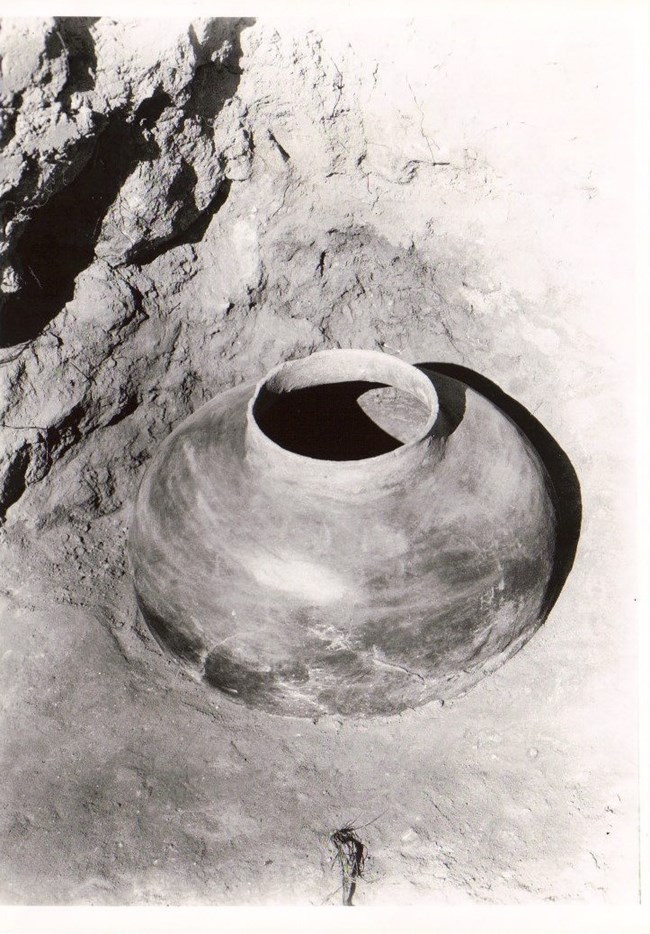 A large olla, a type of clay pot, partially buried in a dirt floor next to a stone wall