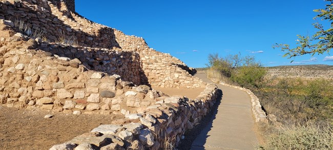A stone pueblo with a paved path alongside it. The path has a slight drop-off on one side and no rails.