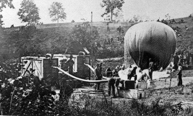 Nine men fill an early war balloon using hoses connected to large wooden wagons