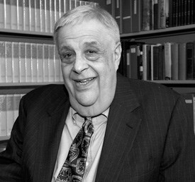 White man in suit and tie sitting in front of a packed bookcase.