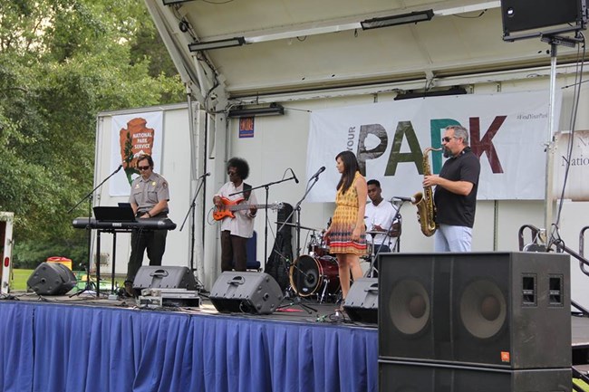 NPS Centennial Band performing at Shiloh National Military Park. Musicians are standing on a stage with a National Park Service banner behind them.