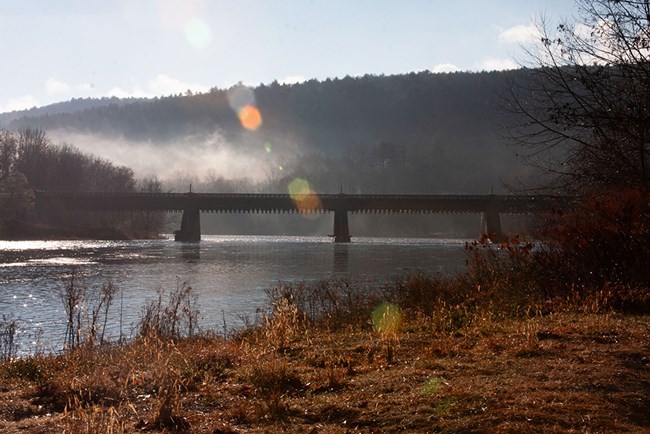 Puente de madera y piedra sobre un río. Al fondo hay colinas con árboles y niebla.