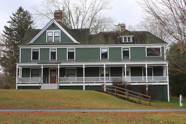 una casa con tres pisos. Es un color gris-verde con adorno blanco y un porche delante de la casa.