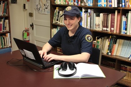 Person wearing volunteer hat and shirt types at a laptop. In front of them on the table is an open notebook and headphones.